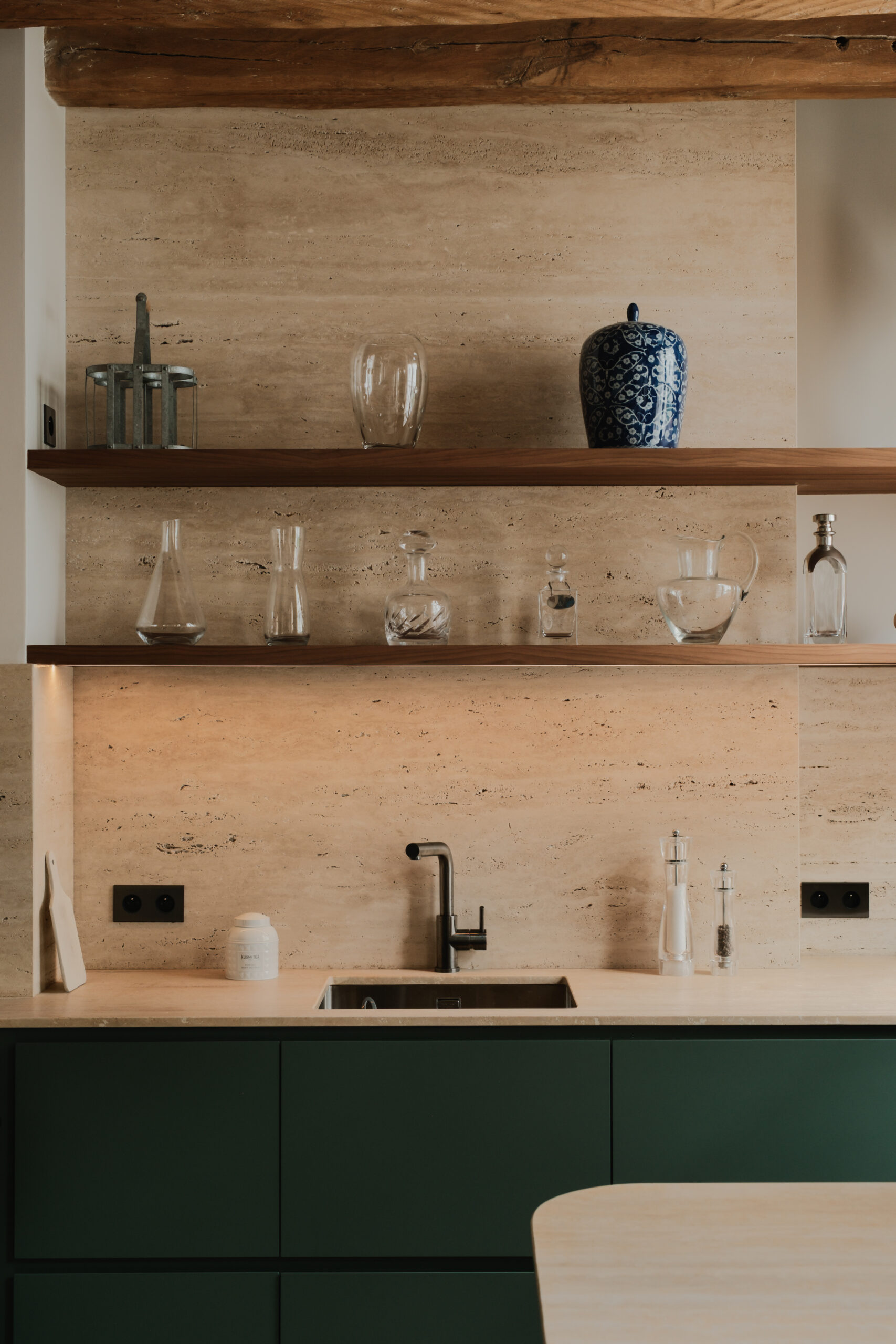 Cuisine sur mesure vert profond, crédence et plan de travail en travertin, étagères ouvertes en noyer, évier intégré, robinetterie contemporaine, matériaux design.