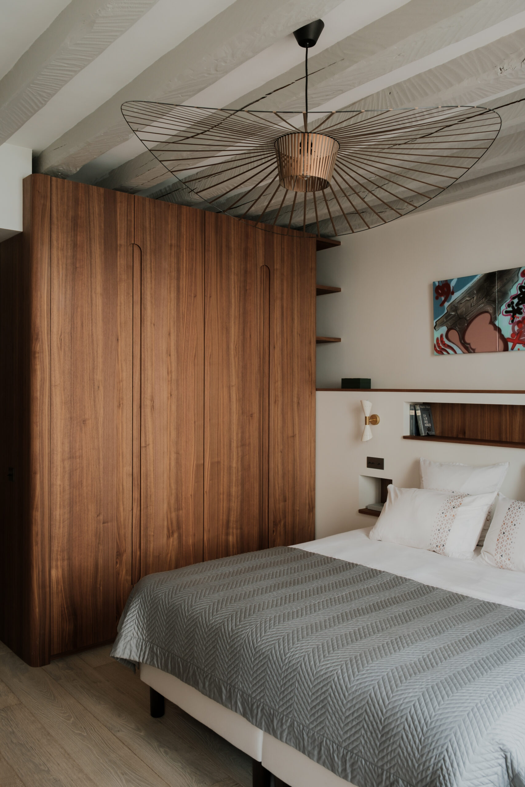 Aménagement de chambre sur mesure, dressing en noyer aux angles arrondis, portes aux poignées intégrées, tête de lit avec niches de rangement, étagères en bois, poutres apparentes, suspension design, matériaux nobles et ambiance chaleureuse.