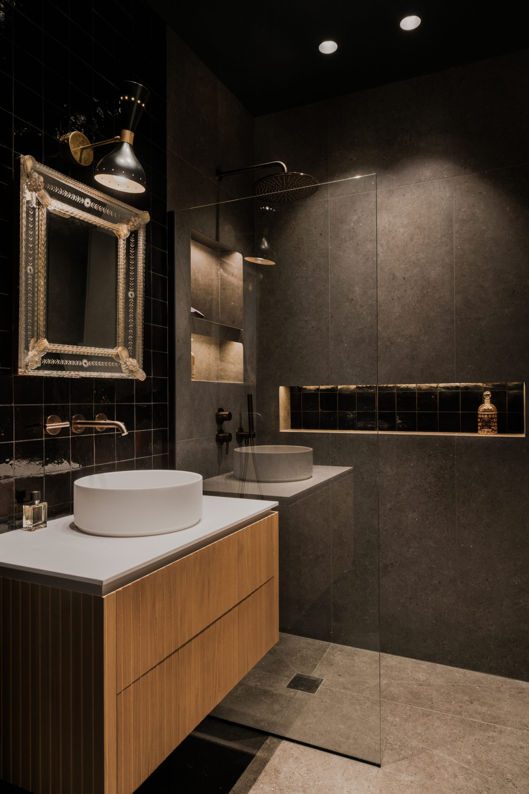 Salle d’eau sur mesure, meuble vasque suspendu en bois cannelé, vasque à poser blanche, douche à l’italienne, carrelage zellige noir brillant, revêtement en pierre grise, niches murales intégrées, robinetterie bronze, design contemporain et ambiance élégante.