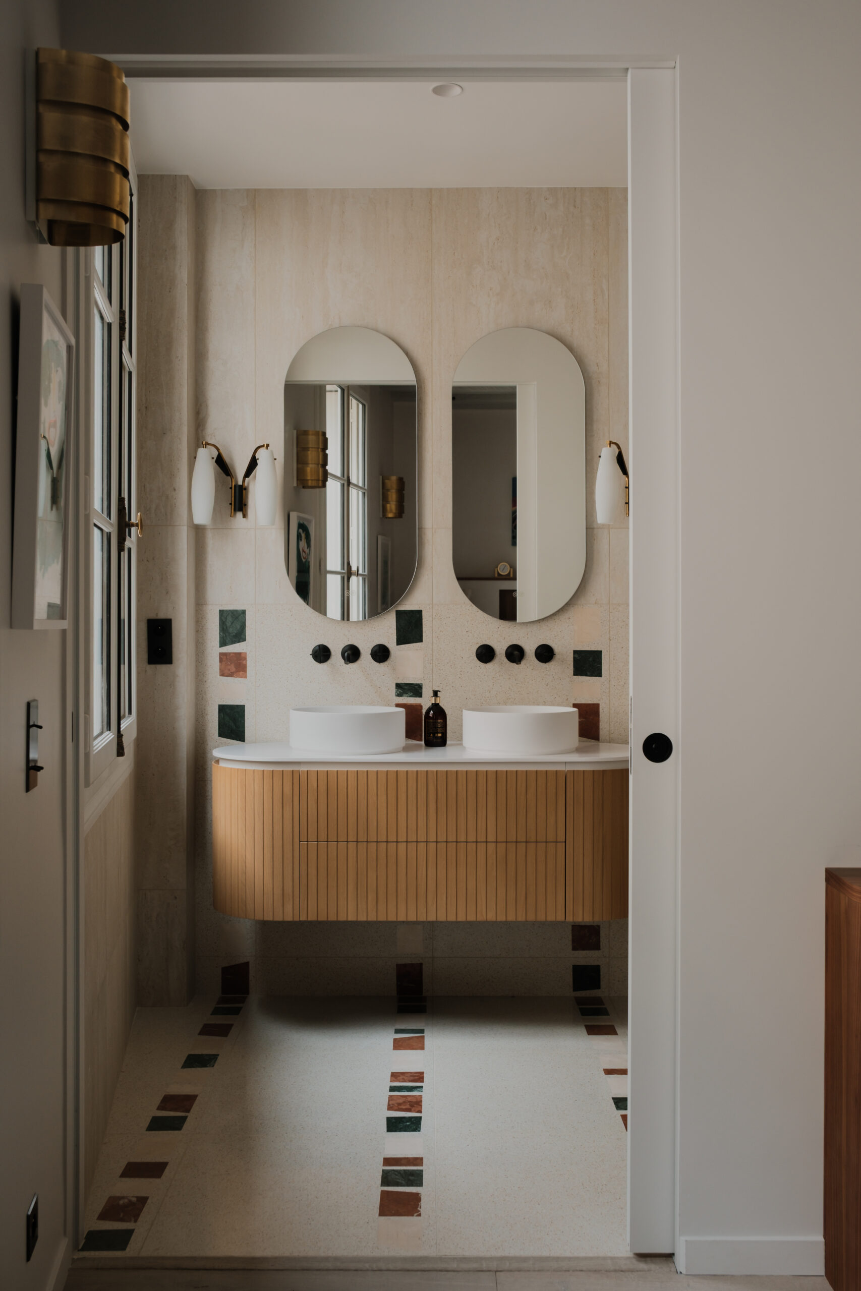 Salle de bain sur mesure, meuble double vasque en bois cannelé aux lignes arrondies, double vasque, miroirs design, robinetterie noire, revêtement effet travertin, détails en marbre coloré, appartement Saint Germain.